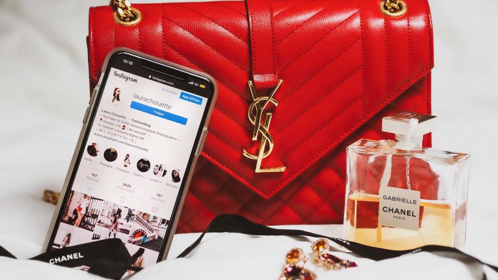 Photo d'un iPhone avec l'application Instagram ouverte où on aperçoit un beau feed de photos. Le téléphone est placé contre un sac de luxe rouge à côté d'une bouteille de parfum.